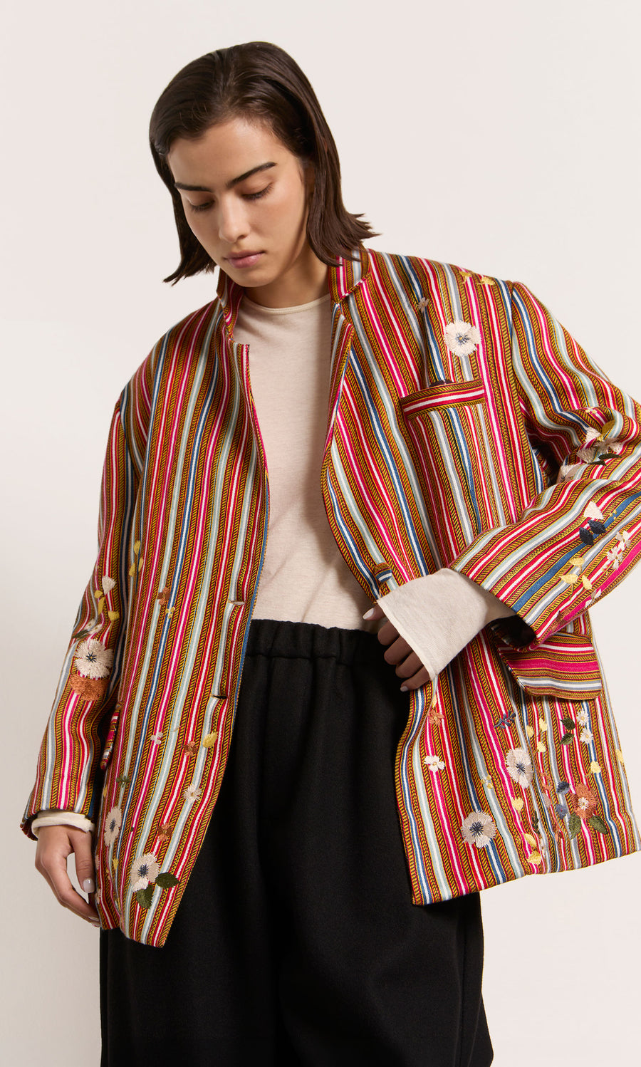 Plümo  Person in a silk blazer with colourful red stripes and hand-stitched flowers, cream shirt, black pants, plain background.