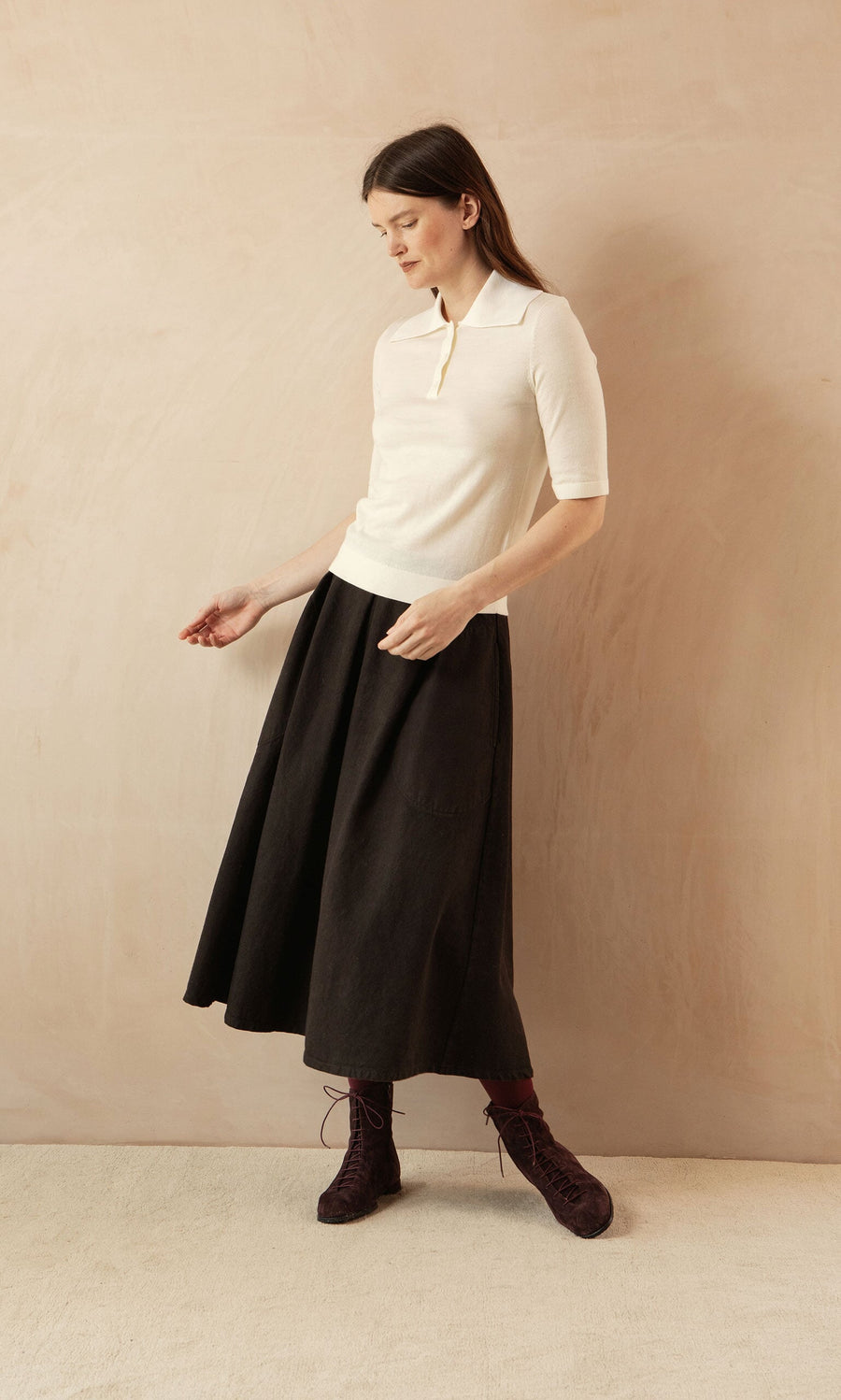 Plümo- Woman wearing a Merino Collar Top and black midi skirt with lace-up boots, posed against a beige background.