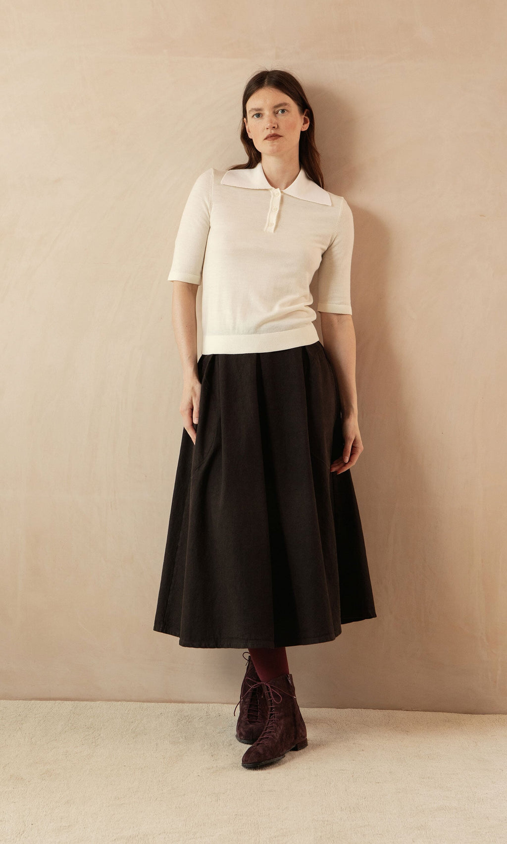 Plümo- Woman in a vintage-inspired look: Merino Collar Top, short sleeves, and long black skirt against a beige wall.