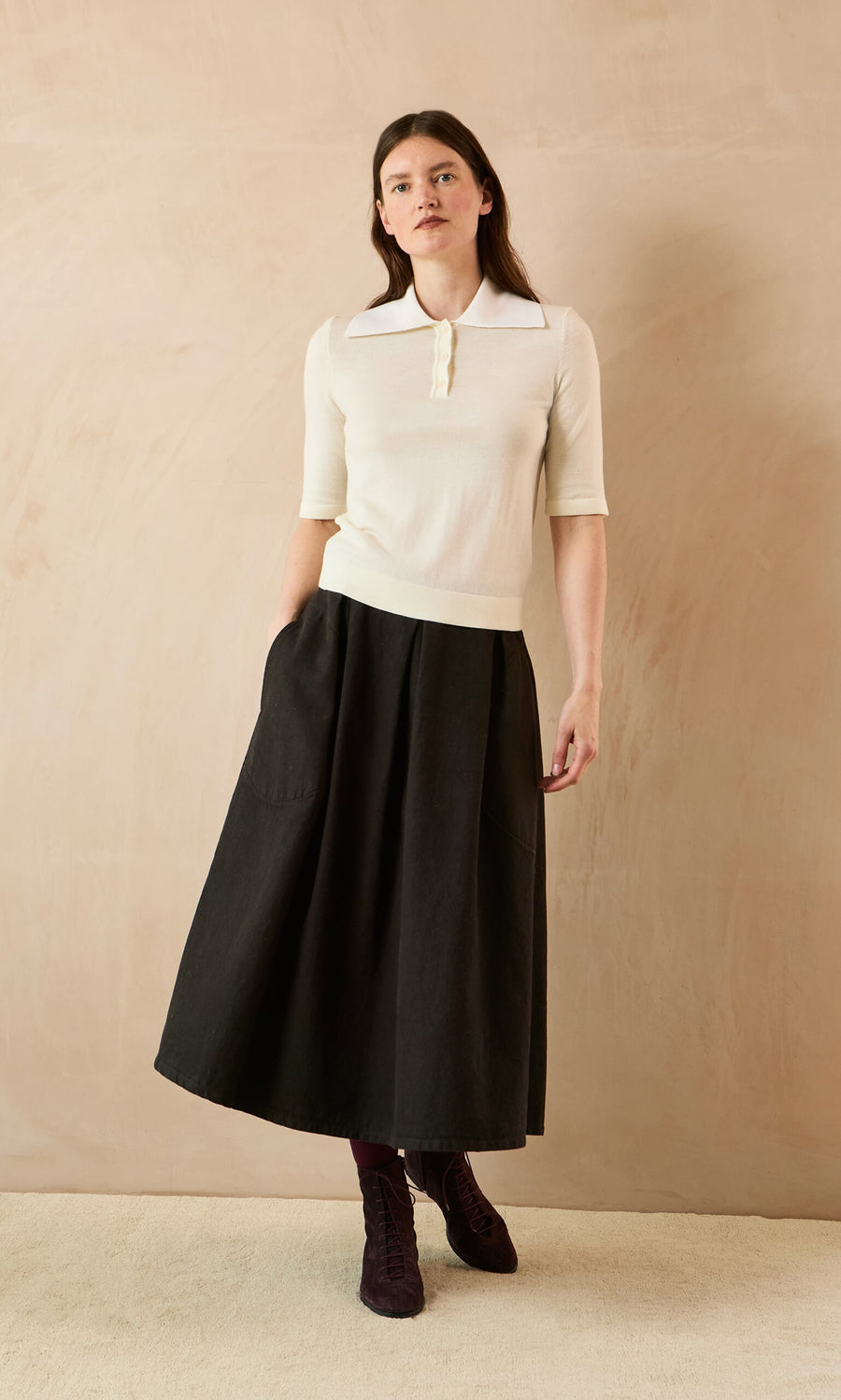 Plümo- Woman in a cream knit top and Allard Skirt stands against a beige background, looking slightly to the side.