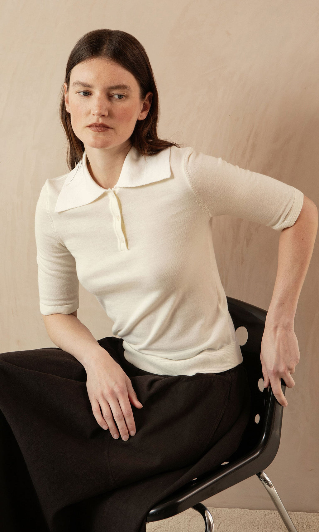 Plümo- Woman with straight brown hair wears the Merino Collar Top and sits on a black chair, looking to the side.