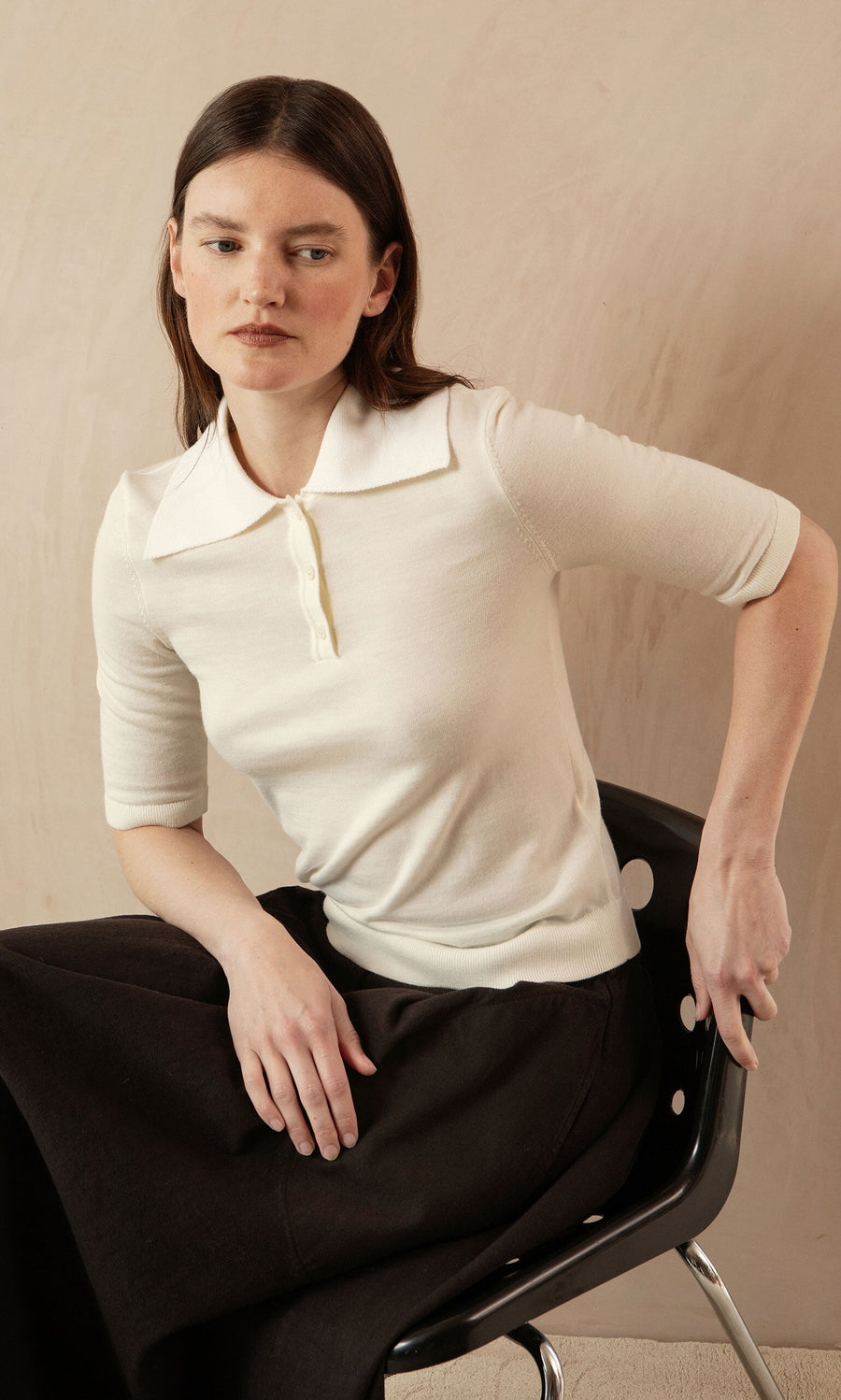 Plümo- Woman with straight brown hair wears the Merino Collar Top and sits on a black chair, looking to the side.