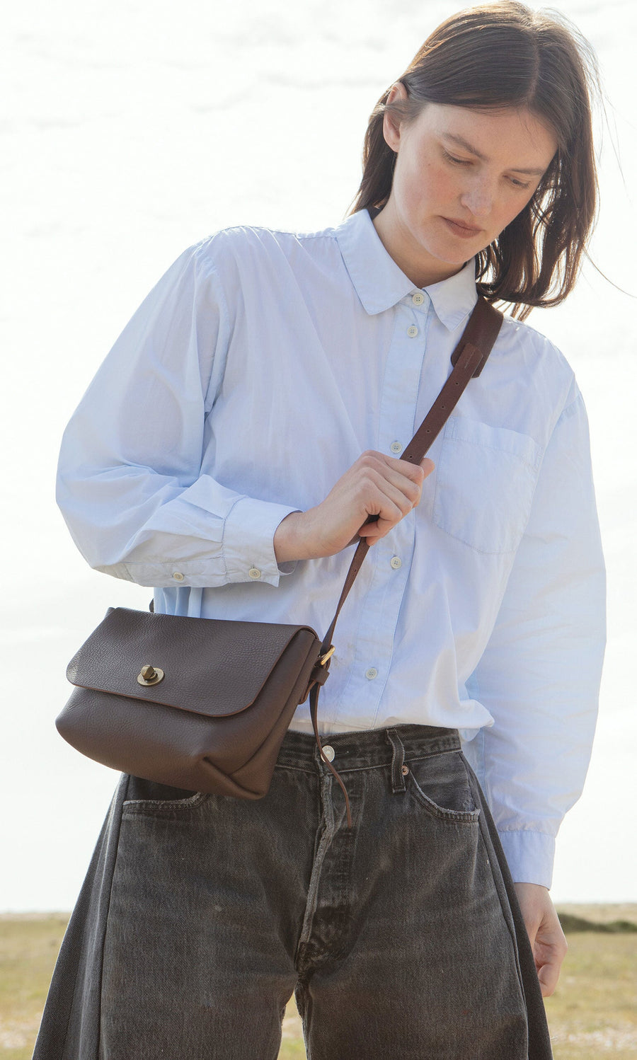 Plümo- Woman in a white button-up and Lasso Jeans - Vintage Black looks down, wearing a brown crossbody bag outdoors.