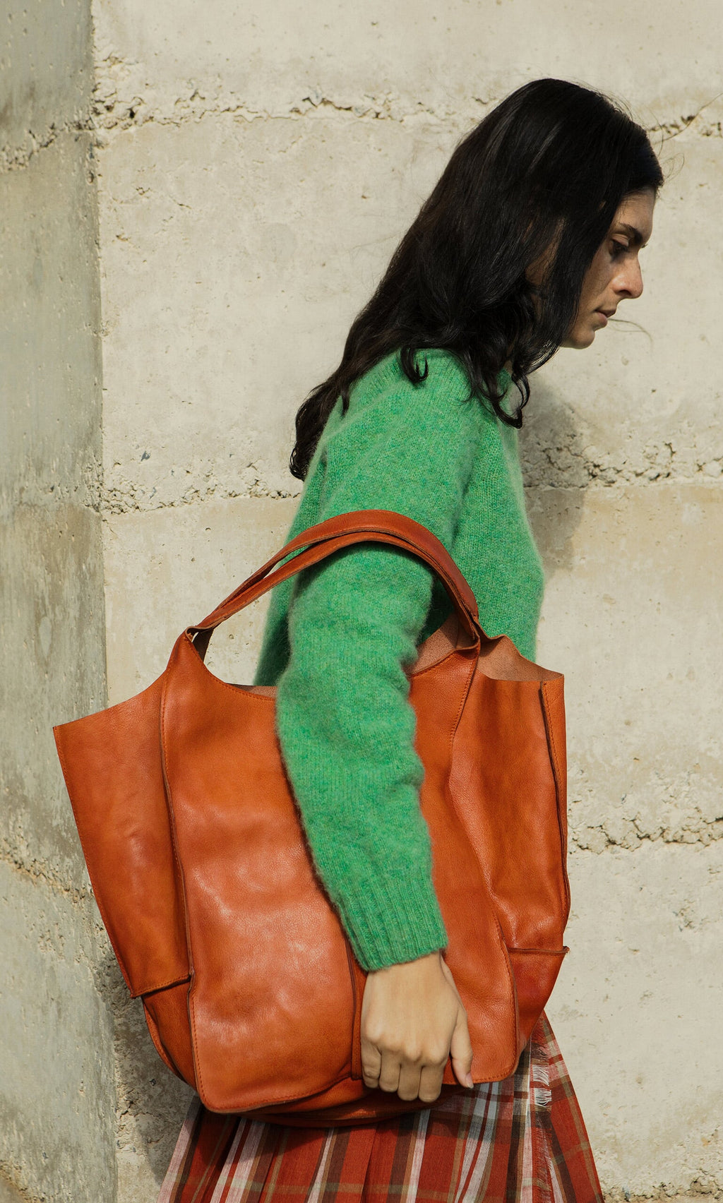 Plümo- Woman in a green sweater carries the Miller Tote, made from vegetable-tanned leather, by a stone wall.
