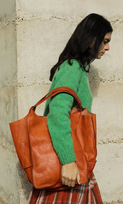 Plümo- Woman in a green sweater carries the Miller Tote, made from vegetable-tanned leather, by a stone wall.