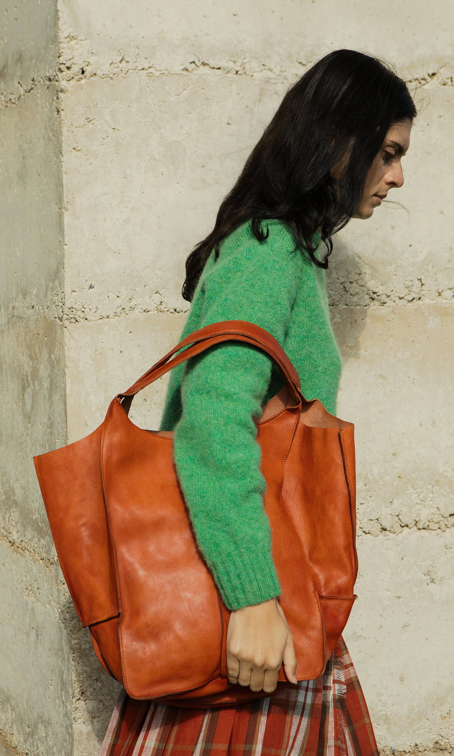 Plümo- Woman in a green sweater carries the Miller Tote, made from vegetable-tanned leather, by a stone wall.