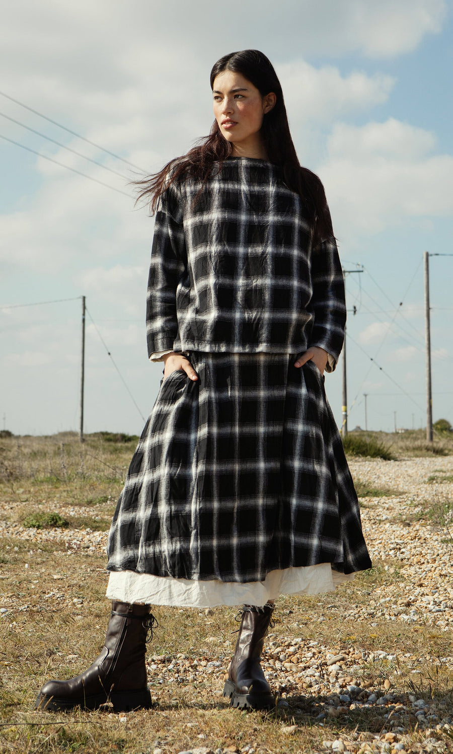 Plümo- A woman in the Gifford Blouse and boots stands outdoors on rocky ground with a cloudy sky behind her.