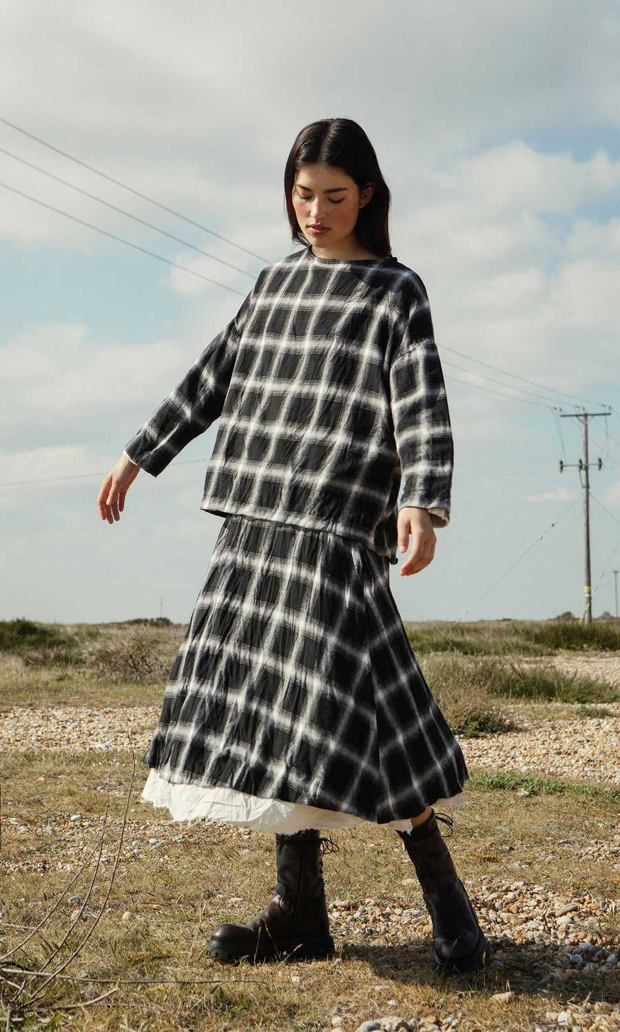 Plümo- A woman wearing the Gifford Blouse and boots stands on grass under a partly cloudy sky.