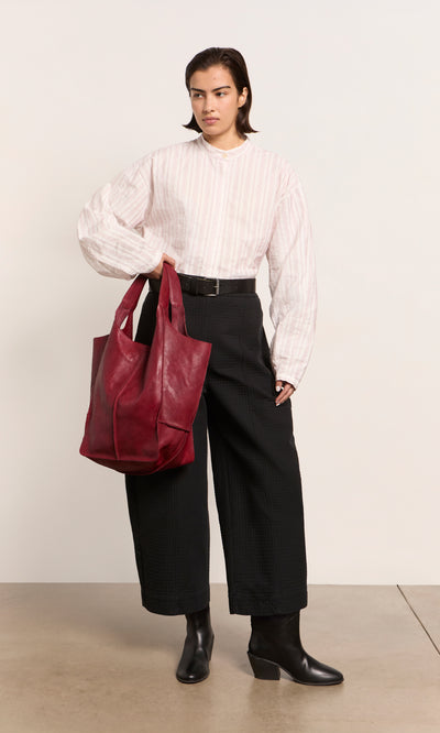 Plümo  Person in a white blouse and black pants holding the Miller Bag in Burnt Sienna, wearing black ankle boots indoors.