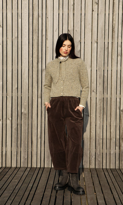 Plümo- Woman stands against a wooden wall in a grey cardigan, brown pants, and artisan-crafted Harrison Boots.