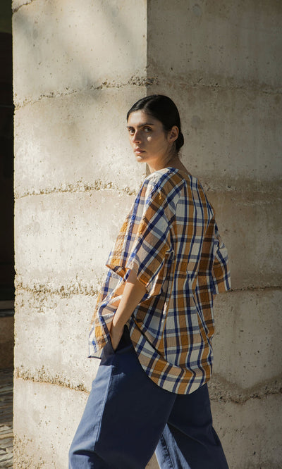 Plümo- A woman in a Blue Madras Shirt and blue pants stands by a concrete pillar, looking over her shoulder in sunlight.