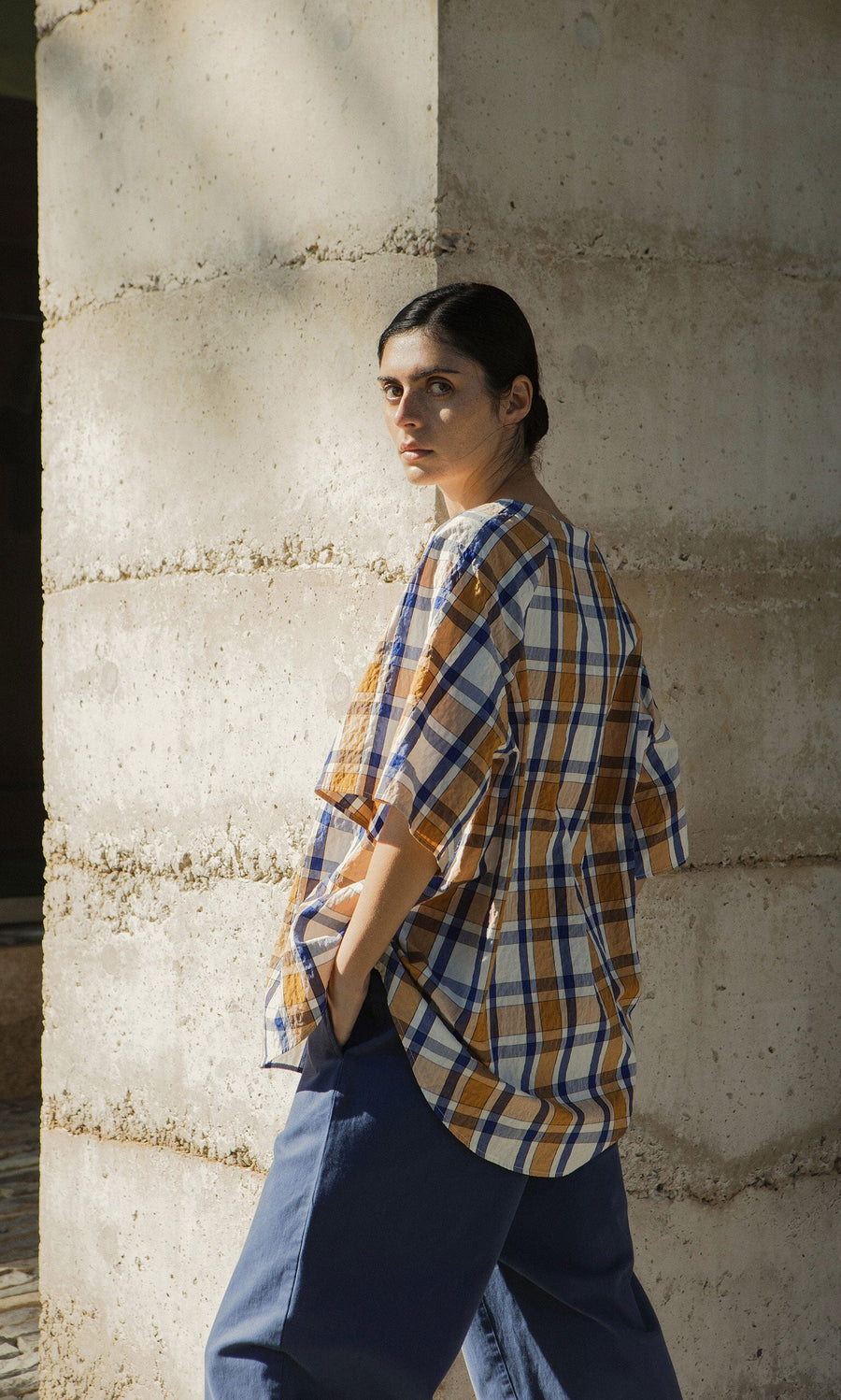 Plümo- A woman in a Blue Madras Shirt and blue pants stands by a concrete pillar, looking over her shoulder in sunlight.