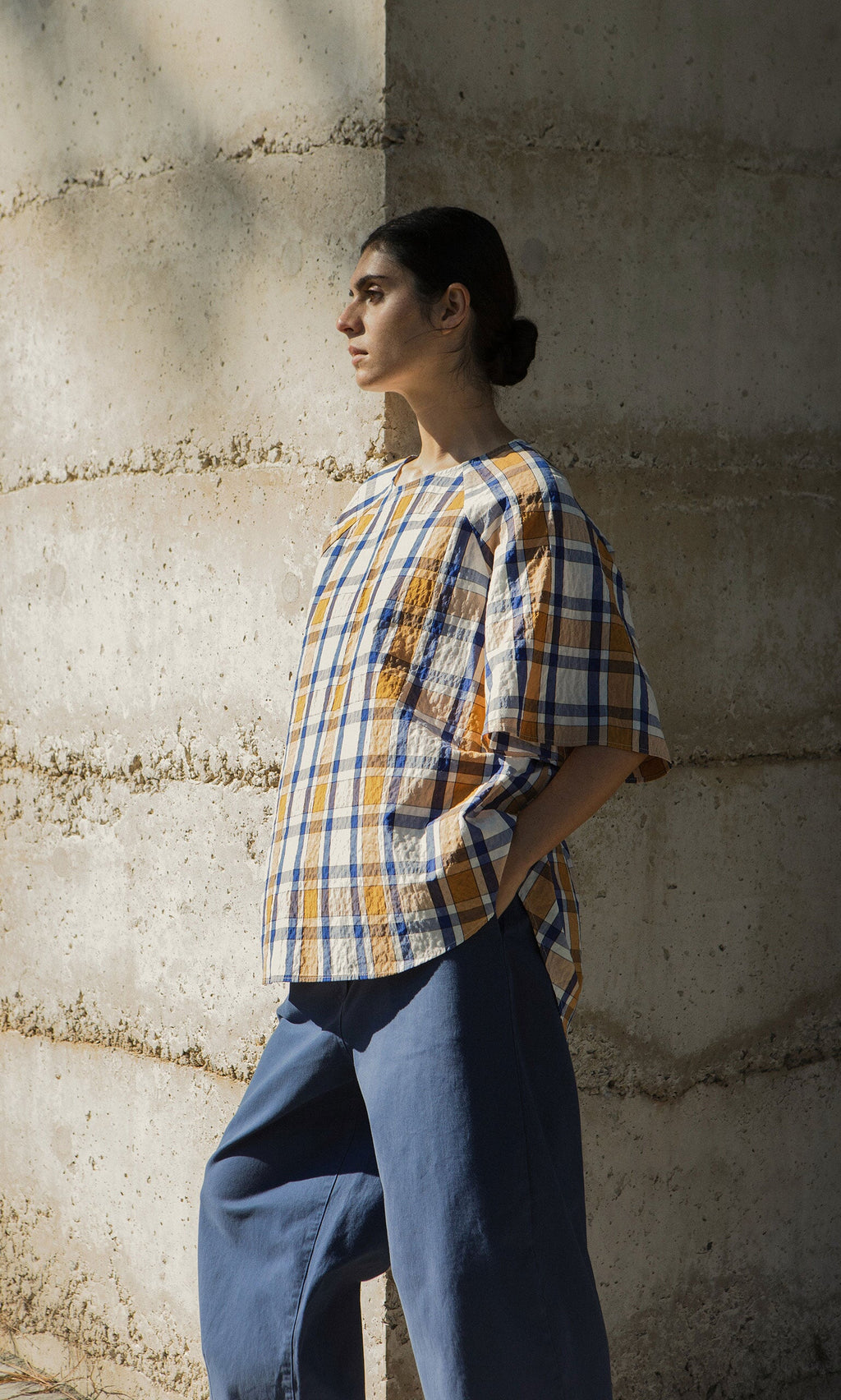 Plümo- Person in a Blue Madras Shirt and blue pants stands against a textured concrete wall, looking left in natural light.