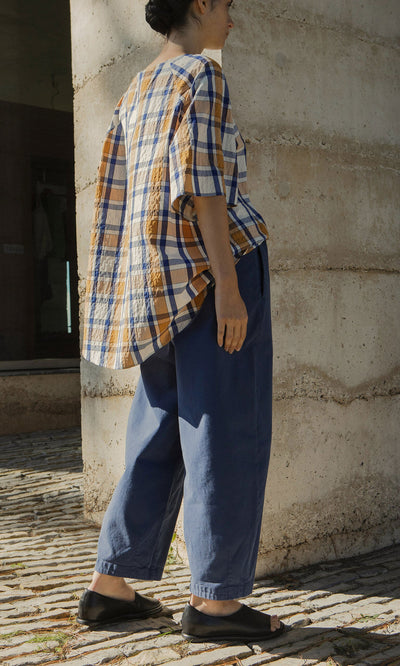 Plümo- Person in Moulder Pants, a plaid shirt, and black sandals standing outdoors by a textured concrete wall.