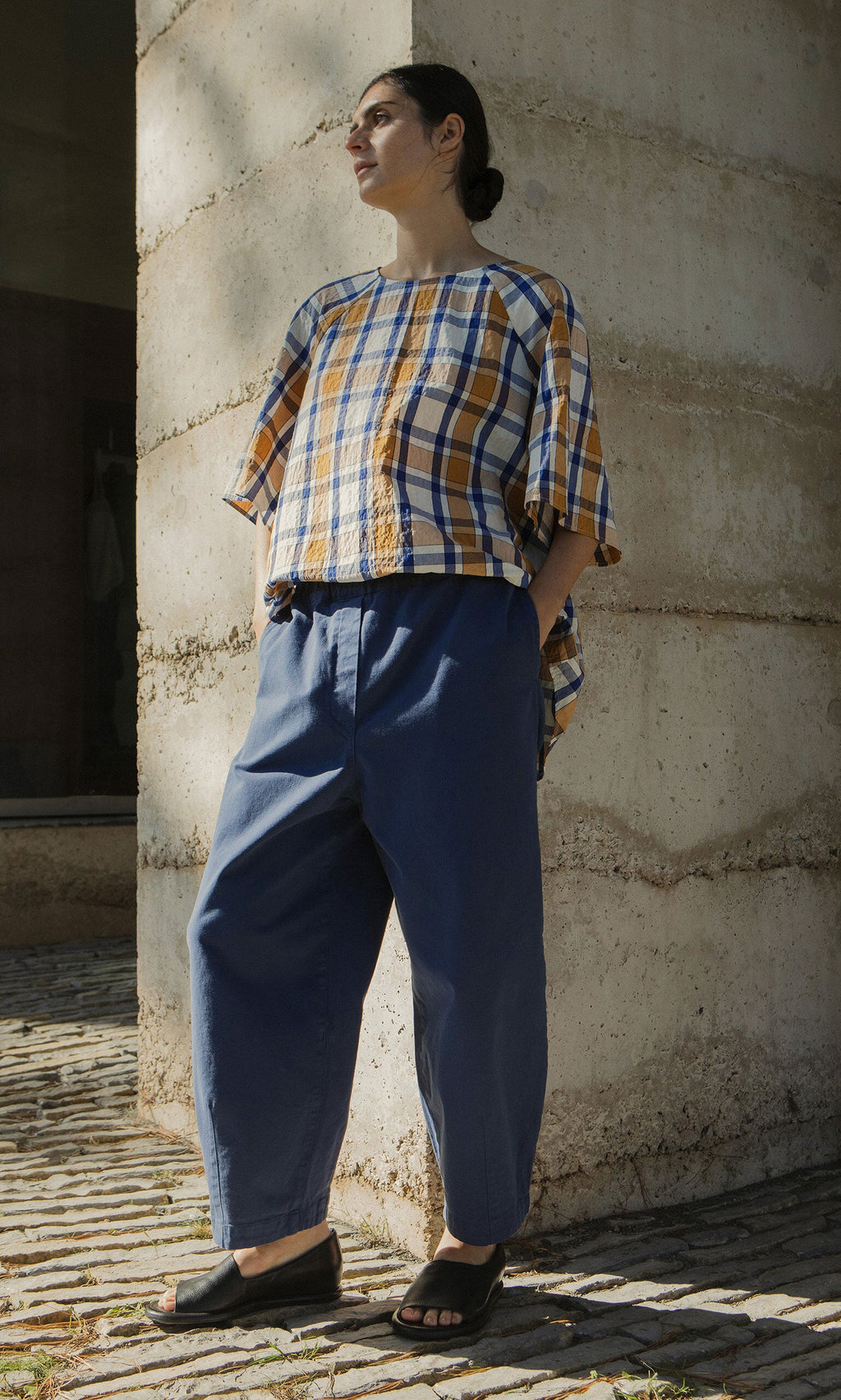 Plümo- A person in Moulder Pants and a plaid top stands outdoors by a concrete wall in sunlight.