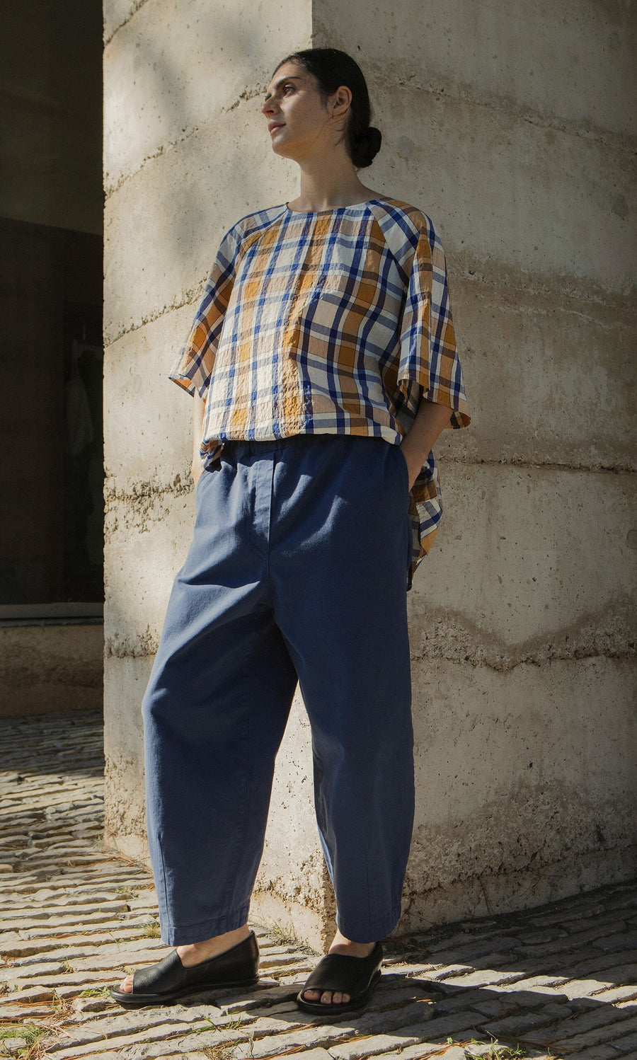 Plümo- A person in Moulder Pants and a plaid top stands outdoors by a concrete wall in sunlight.