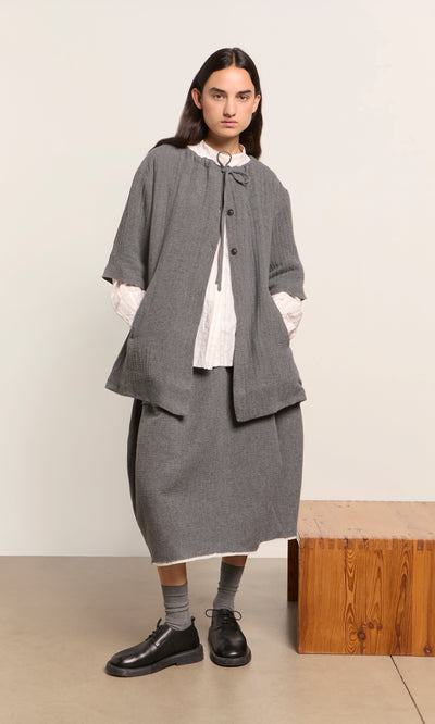 Plümo  Someone in a Moy Jacket and gray skirt stands by a wooden bench, hands in pockets, against a plain background.