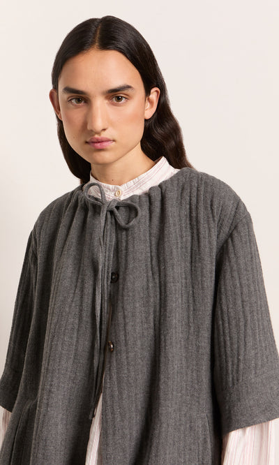 Plümo  Person with long dark hair in the Moy Jacket over a light collared shirt, looking at the camera.