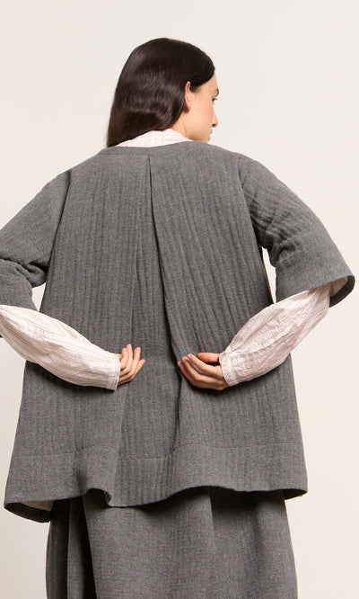 Plümo  Woman in the Moy Jacket, grey pleated-textured wool, white shirt, seen from behind with hands clasped.