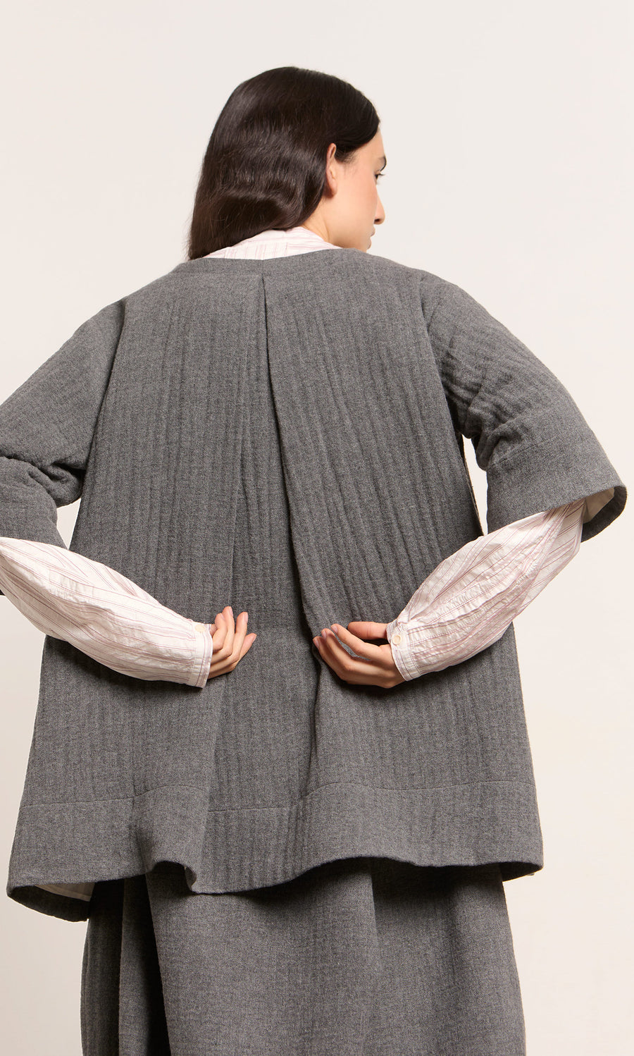 Plümo  Woman in the Moy Jacket, grey pleated-textured wool, white shirt, seen from behind with hands clasped.