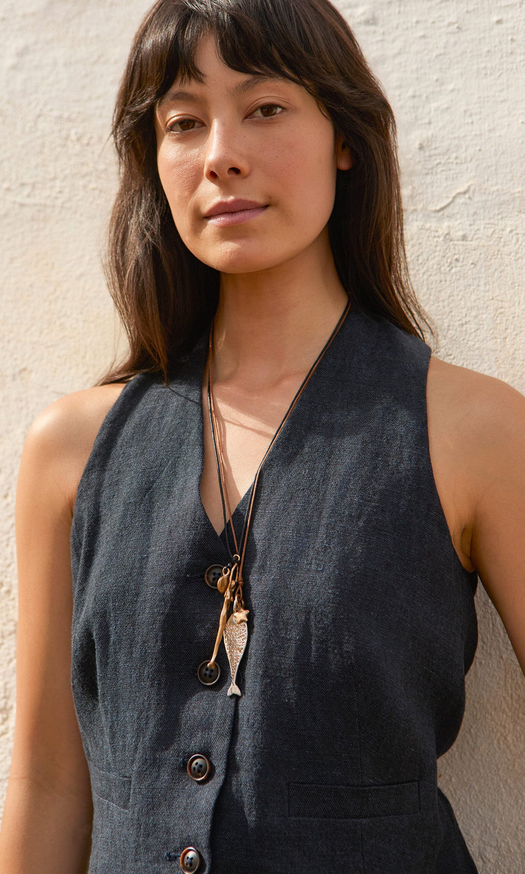 Plümo- A woman in a sleeveless dark vest stands against a textured wall, wearing layered necklaces, including the Capri Necklace.
