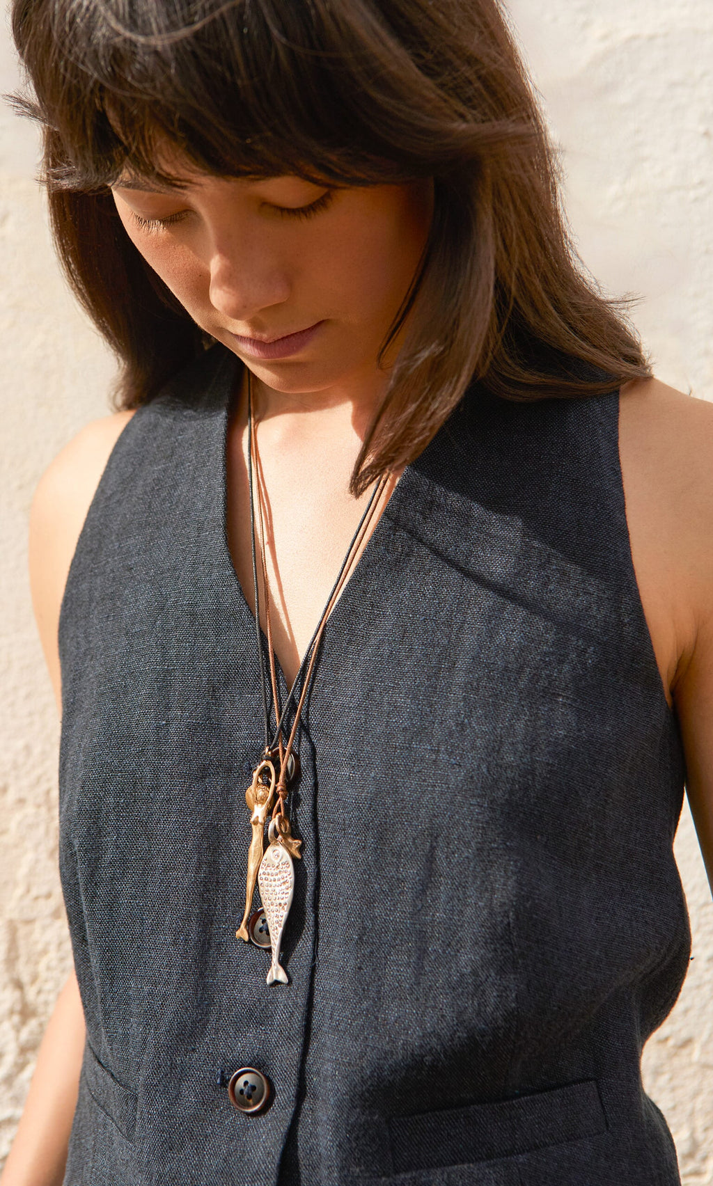 Plümo- A woman in a sleeveless dark vest looks down, wearing layered Capri Necklaces with ocean charm and fish pendants.