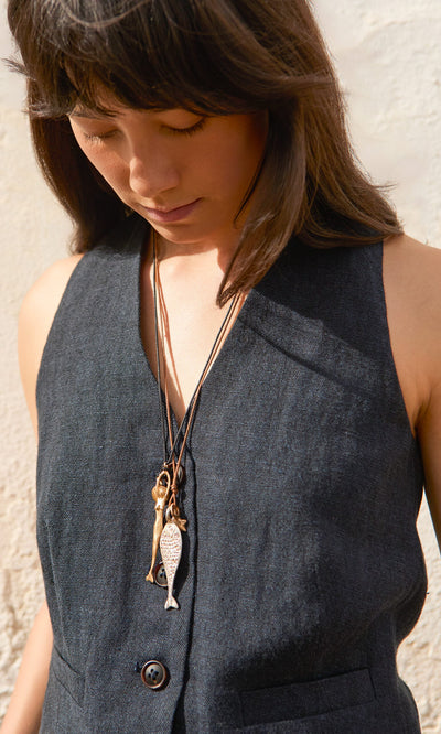 Plümo- A woman in a sleeveless dark vest looks down, wearing layered Capri Necklaces with ocean charm and fish pendants.