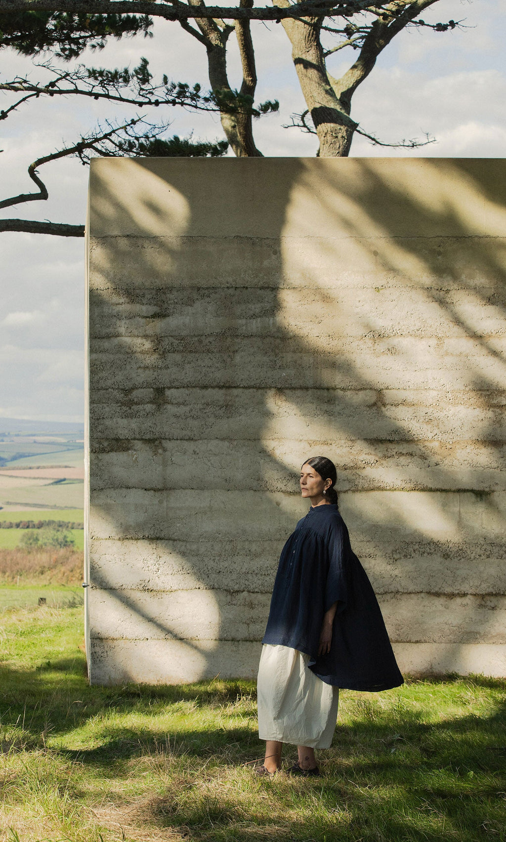 Plümo- A woman in the Thistle Blouse - Deep Navy stands by a textured concrete wall under dappled sunlight and trees.