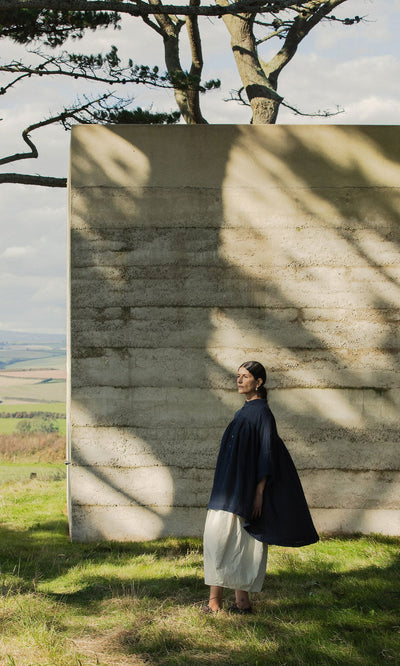 Plümo- A woman in the Thistle Blouse - Deep Navy stands by a textured concrete wall under dappled sunlight and trees.