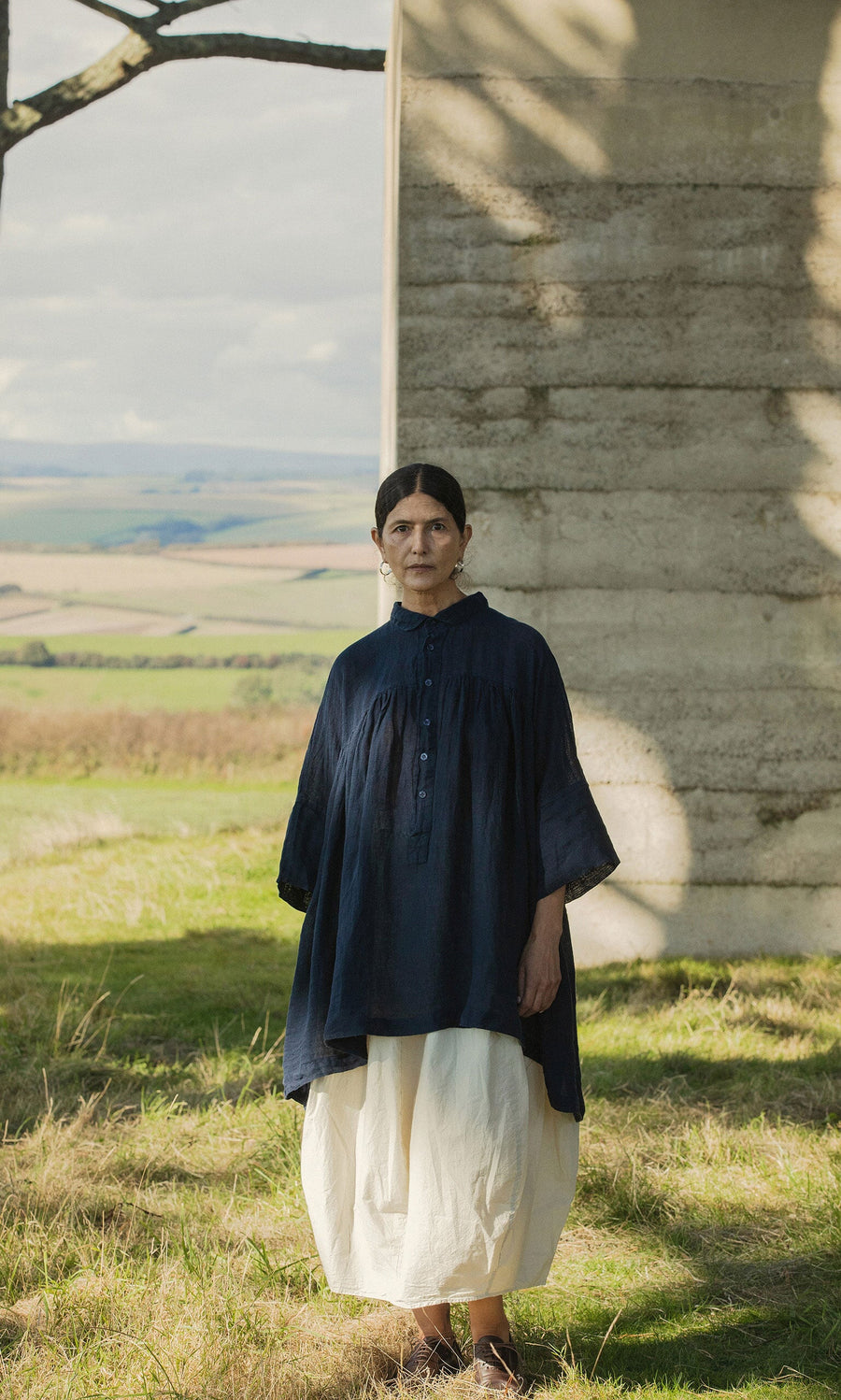 Plümo- A woman in the Thistle Blouse - Deep Navy with cropped sleeves stands by concrete, fields stretching out behind her.