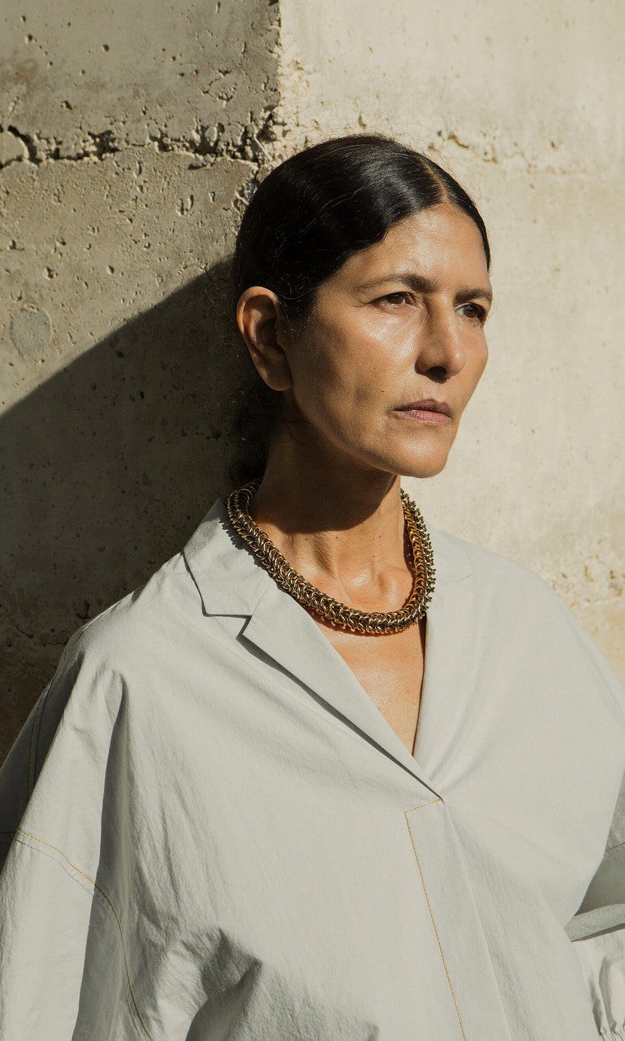 Plümo- Woman with dark hair in a light shirt wears the Kibbin Necklace, standing against a textured stone wall in sunlight.