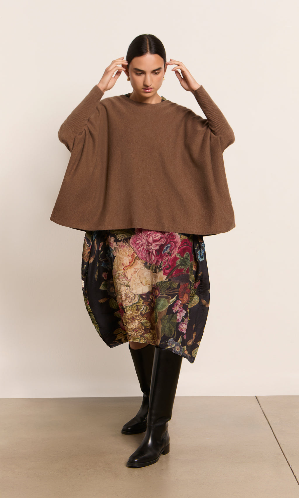 Plümo  Woman in a brown Neves Crew Neck sweater, floral skirt, and black knee-high boots stands against a plain wall.