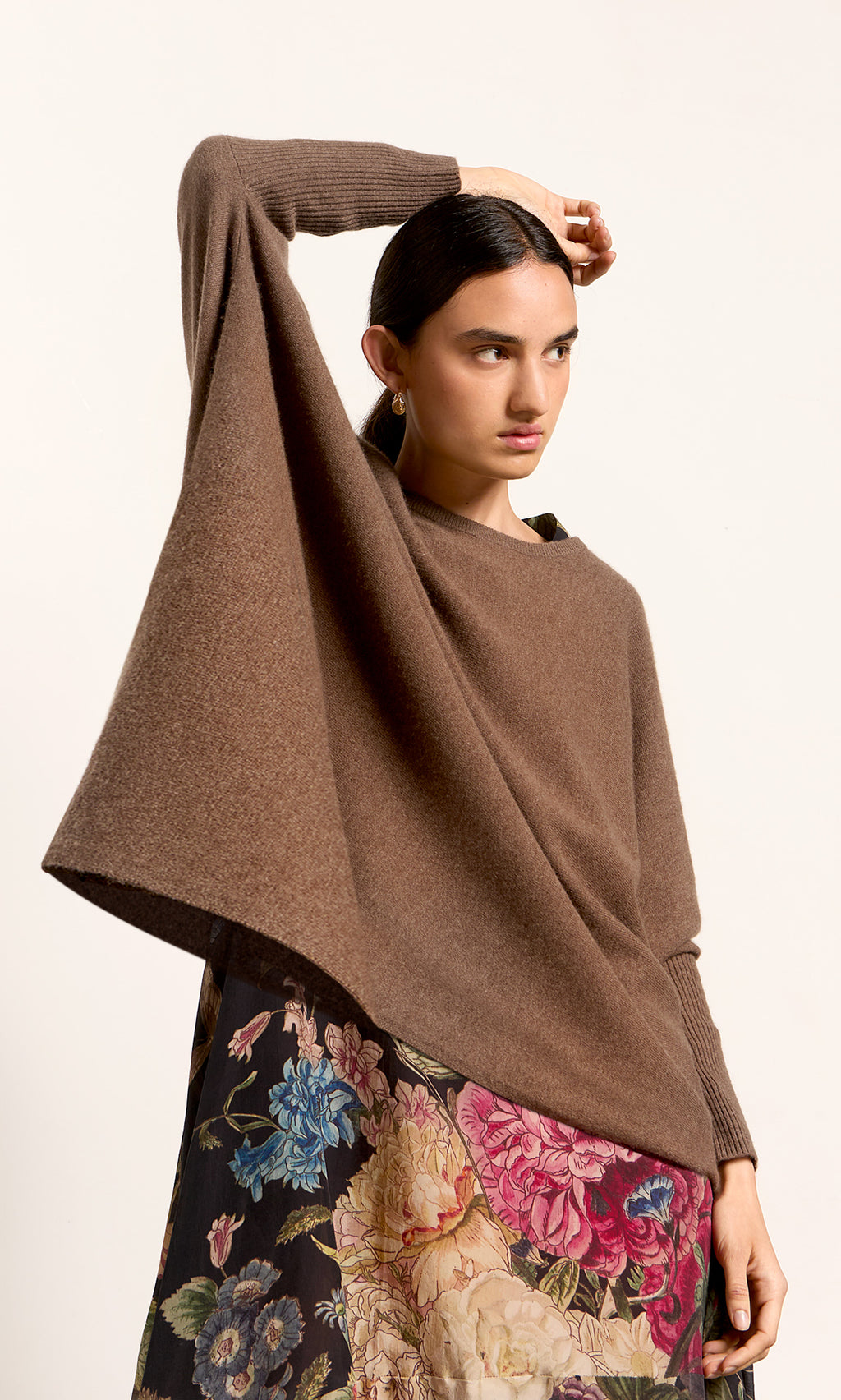 Plümo  Woman in a cumin Neves Crew Neck wool jumper and floral skirt, posing with one arm raised over her head.