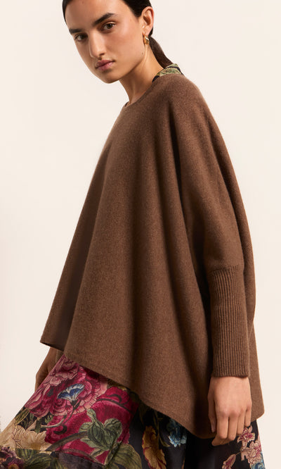 Plümo  Woman in a cumin Neves Crew Neck sweater and floral skirt stands against a plain background, looking at the camera.