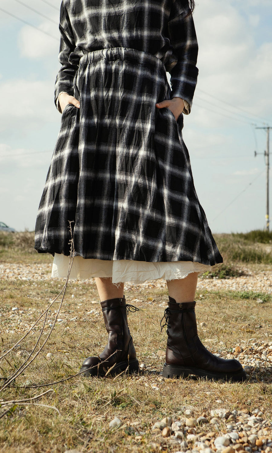 Plümo- Person in the Noemi Skirt and black boots stands on rocky ground with hands in pockets.