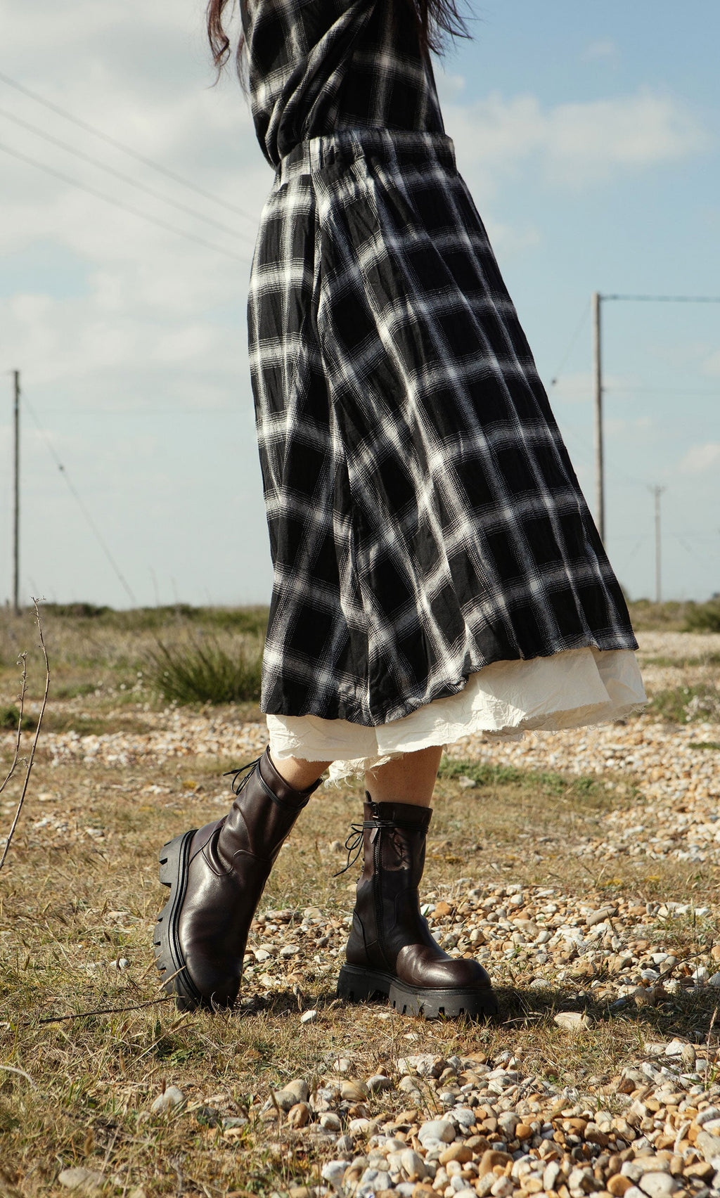 Plümo- A person in the Noemi Skirt and black boots walks on a rocky, grassy field under a partly cloudy sky.