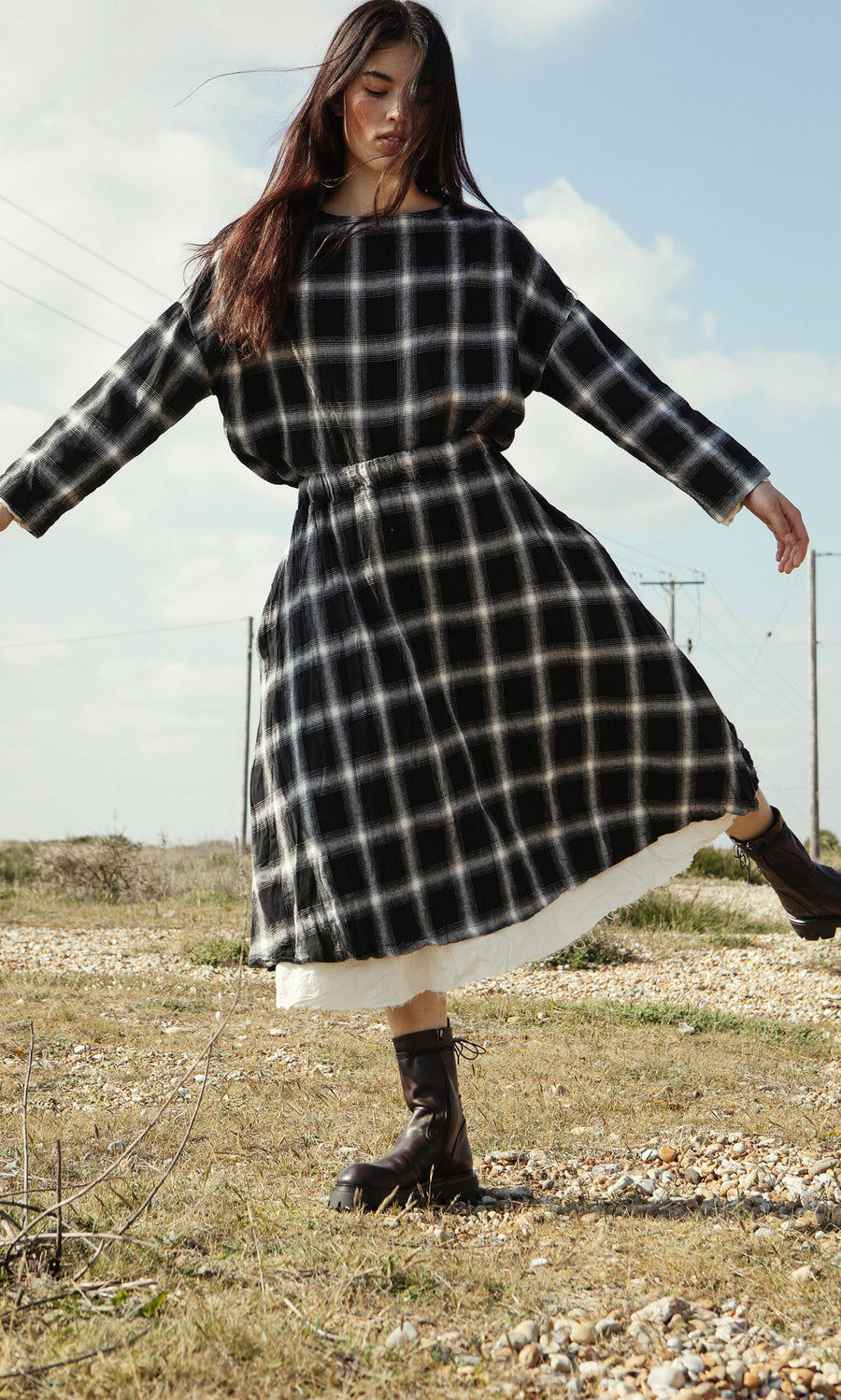 Plümo- Woman in the Noemi Skirt and boots balances on one foot outdoors in a grassy, rocky field under a blue sky.