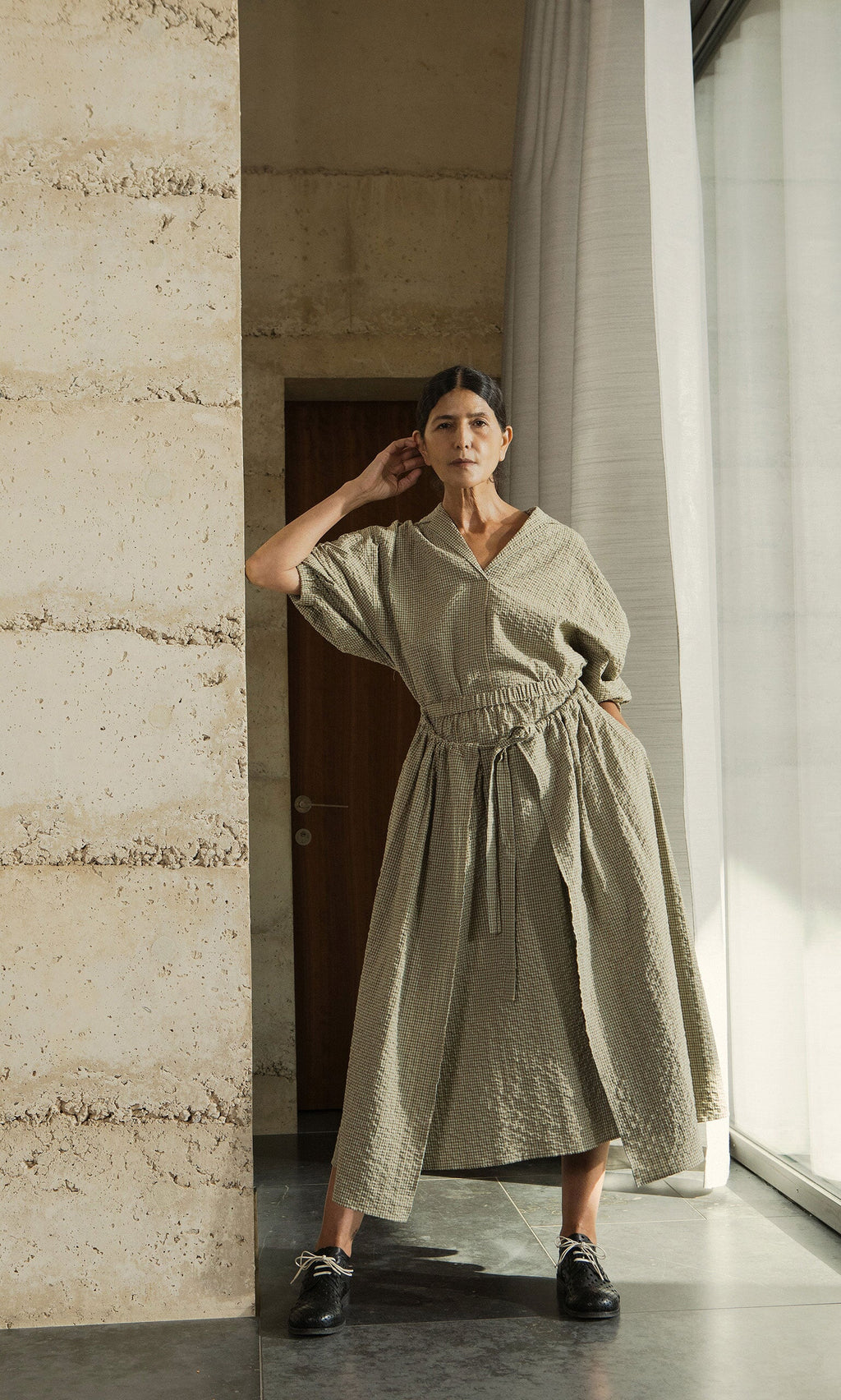 Plümo- Woman wears the Fern Blouse in textured light beige cotton, posing confidently indoors by a stone wall and window.