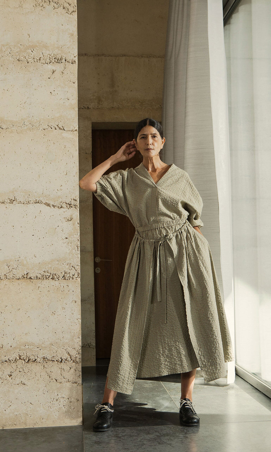 Plümo- Woman wears the Fern Blouse in textured light beige cotton, posing confidently indoors by a stone wall and window.