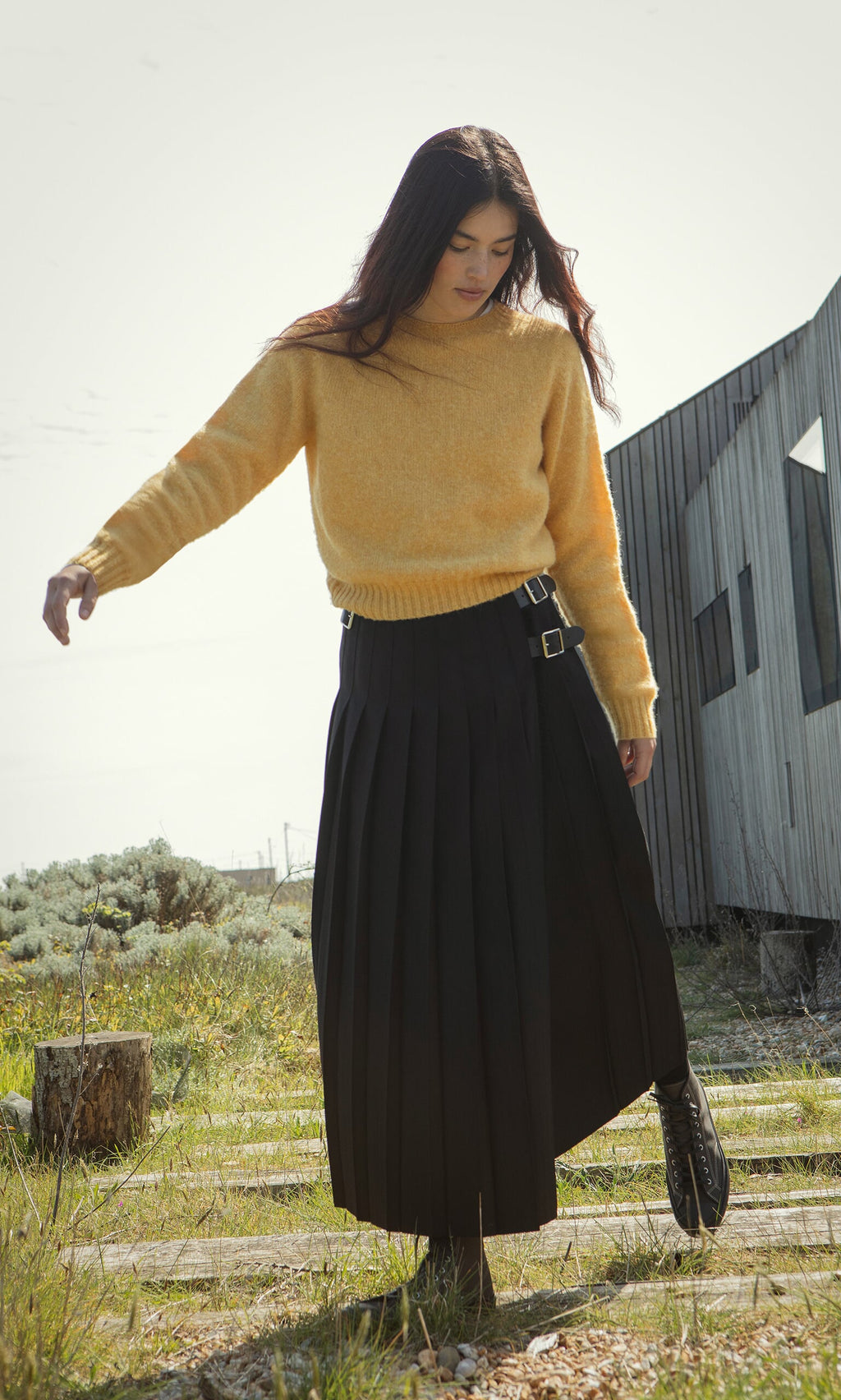 Plümo- A woman in the boxy fit Oban Sweater and a black pleated skirt stands outdoors on wooden planks near a modern building.