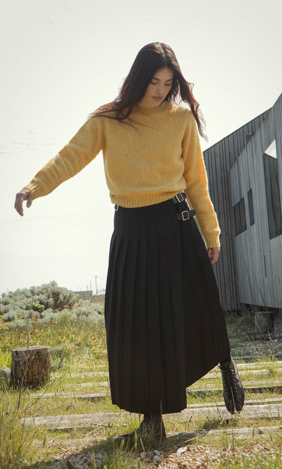 Plümo- A woman in the boxy fit Oban Sweater and a black pleated skirt stands outdoors on wooden planks near a modern building.