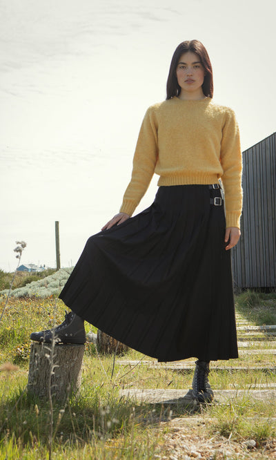 Plümo- Woman outdoors in a boxy fit Oban Sweater and black pleated skirt, holding her skirt with one hand.