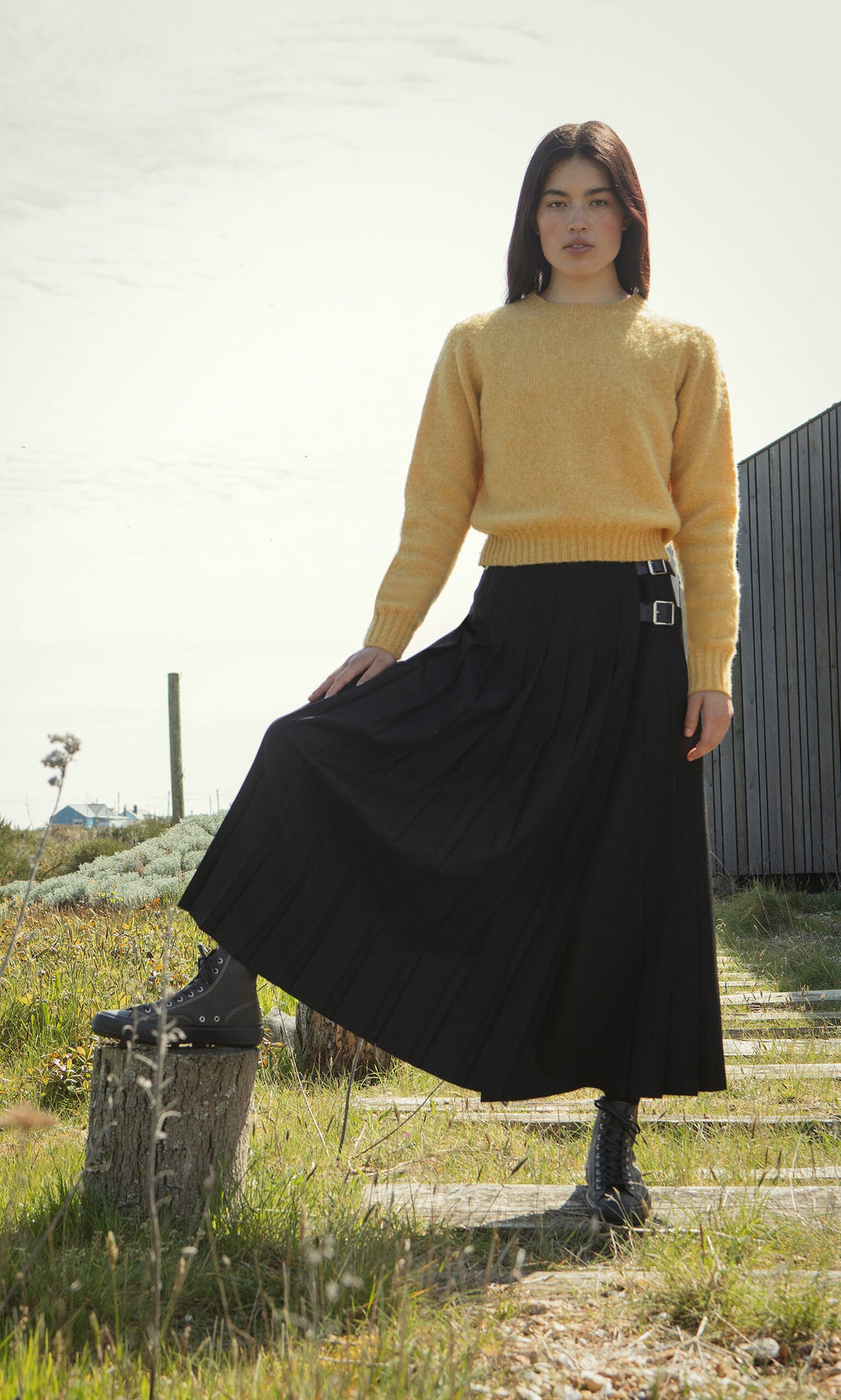 Plümo- Woman in a yellow sweater and Storm Kilt stands outdoors, one foot on a tree stump, looking at the camera.