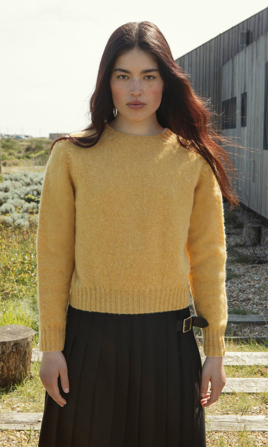 Plümo- Woman with long dark hair wears the Oban Sweater, a brushed wool jumper with boxy fit, by a modern building.