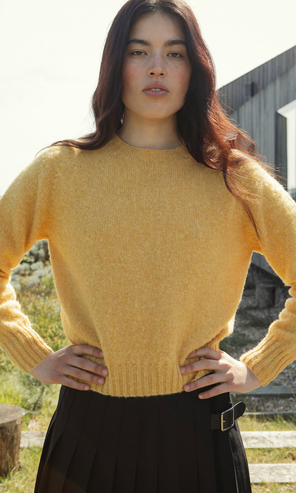 Plümo- Woman with long dark hair wears the Oban Sweater, a boxy yellow jumper, and a black skirt outdoors, hands on hips.
