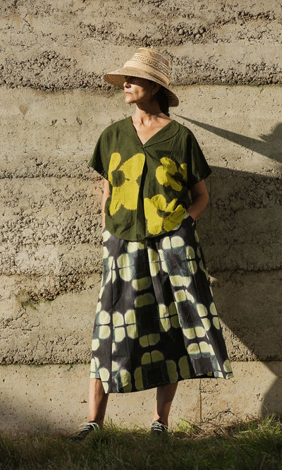 Plümo- Woman in a Bogota Hat and patterned green outfit stands against a textured concrete wall, hands in skirt pockets.