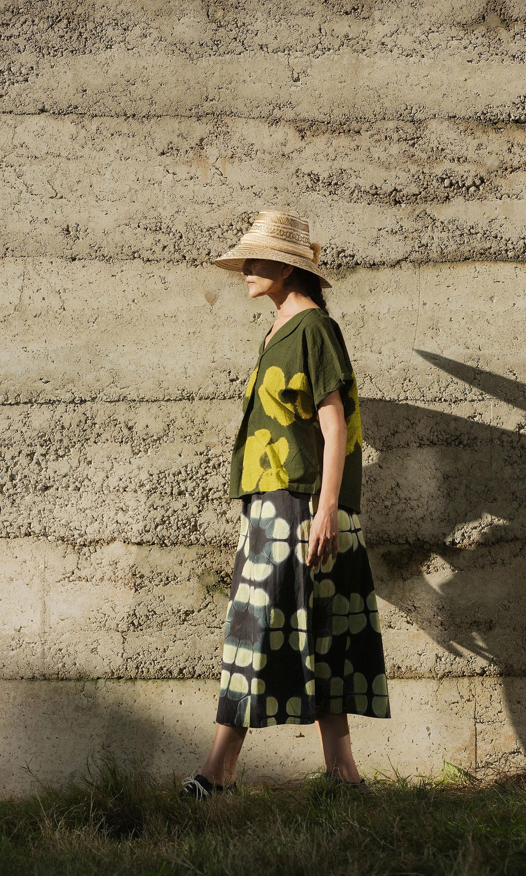 Plümo- A person in a straw hat and green Orla Flower Skirt walks by a rough concrete wall in sunlight.