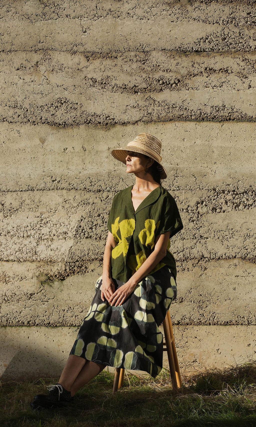 Plümo- A woman in the Orla Flower Skirt and a hat sits on a stool by a sunlit textured concrete wall.