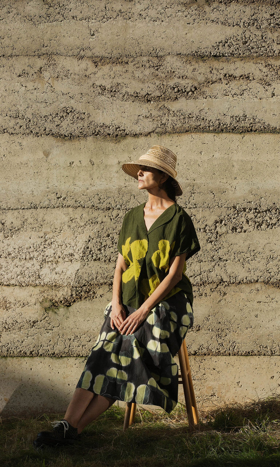 Plümo- A woman in the Orla Flower Skirt and a hat sits on a stool by a sunlit textured concrete wall.