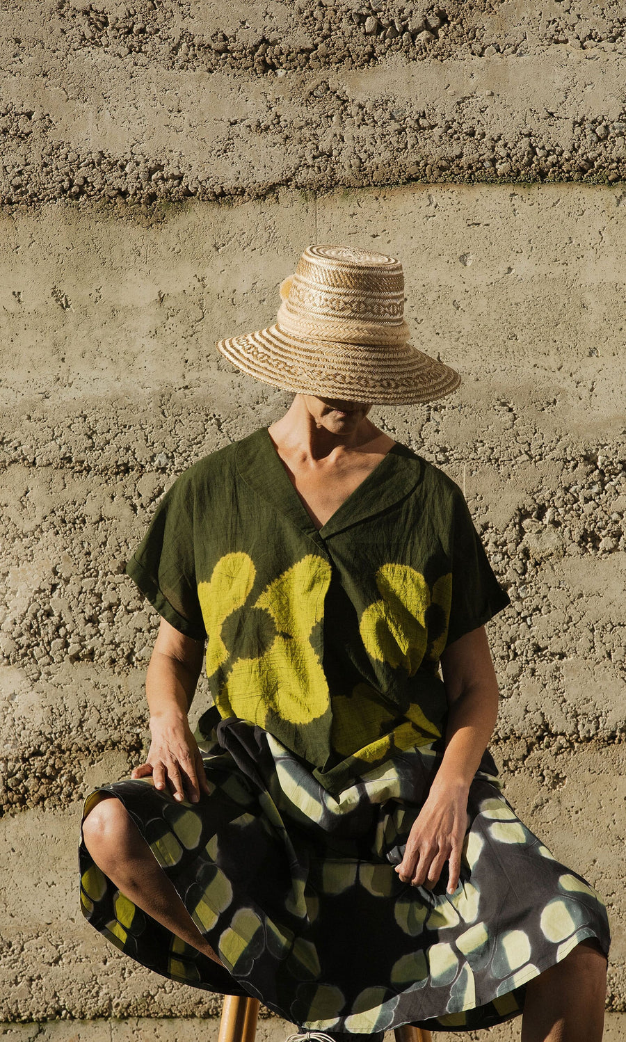 Plümo- A person in a Bogota Hat and patterned dress sits against a textured, striped concrete wall.